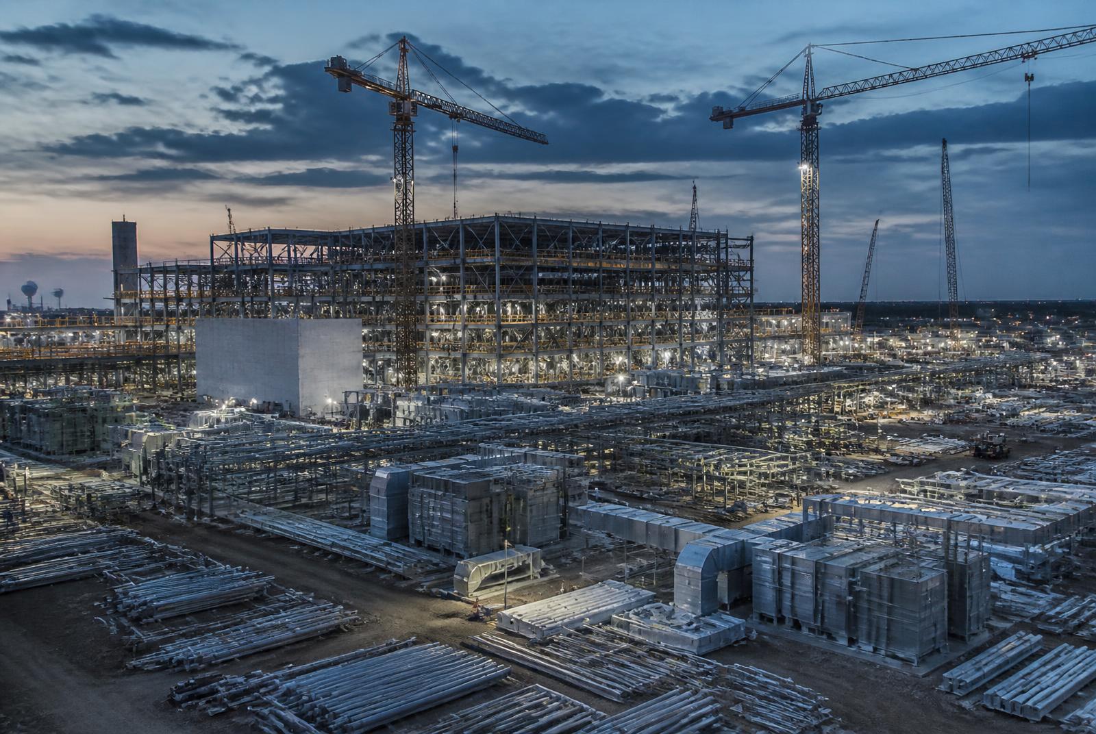Large advanced-manufacturing plant under construction at dusk with structural steel, tower cranes and staged mechanical equipment